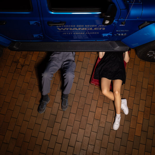 Mechaniker und Kundin liegen unter dem Auto zur Begutachtung des Problems, Blick von oben. Schriftzug: Entdecke den neuen Jeep Wrangler. Jetzt probefahren.